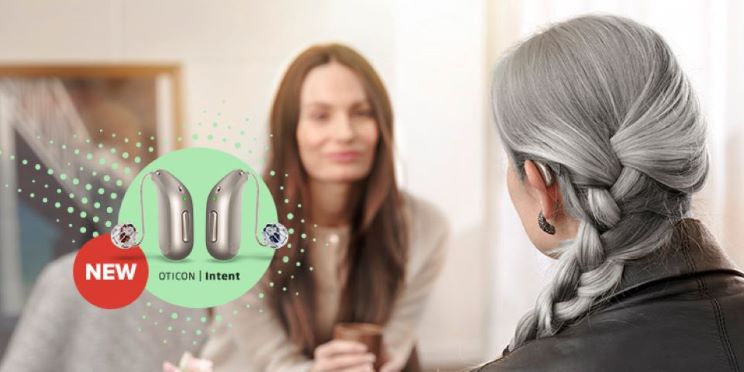 Behind-the-ear hearing aids labeled as 'Oticon Intent' with a red 'New' badge, displayed in a conversation setting with two women, one with gray braided hair in the foreground.